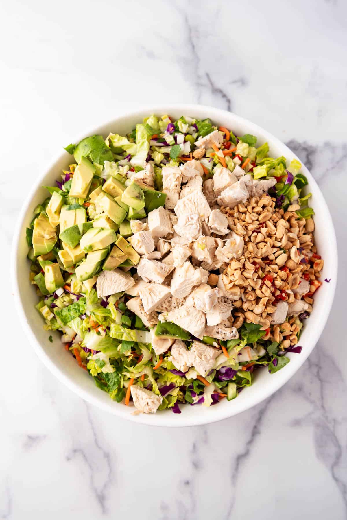 Adding chopped cooked chicken, chopped peanuts, and diced avocado to a bowl of salad.