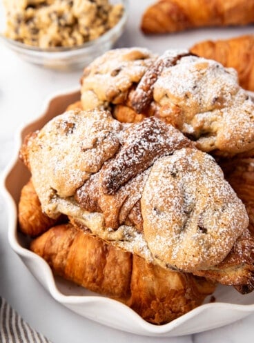 An image of cookie croissants stacked on flaky croissants with a bowl of cookie dough in the background.