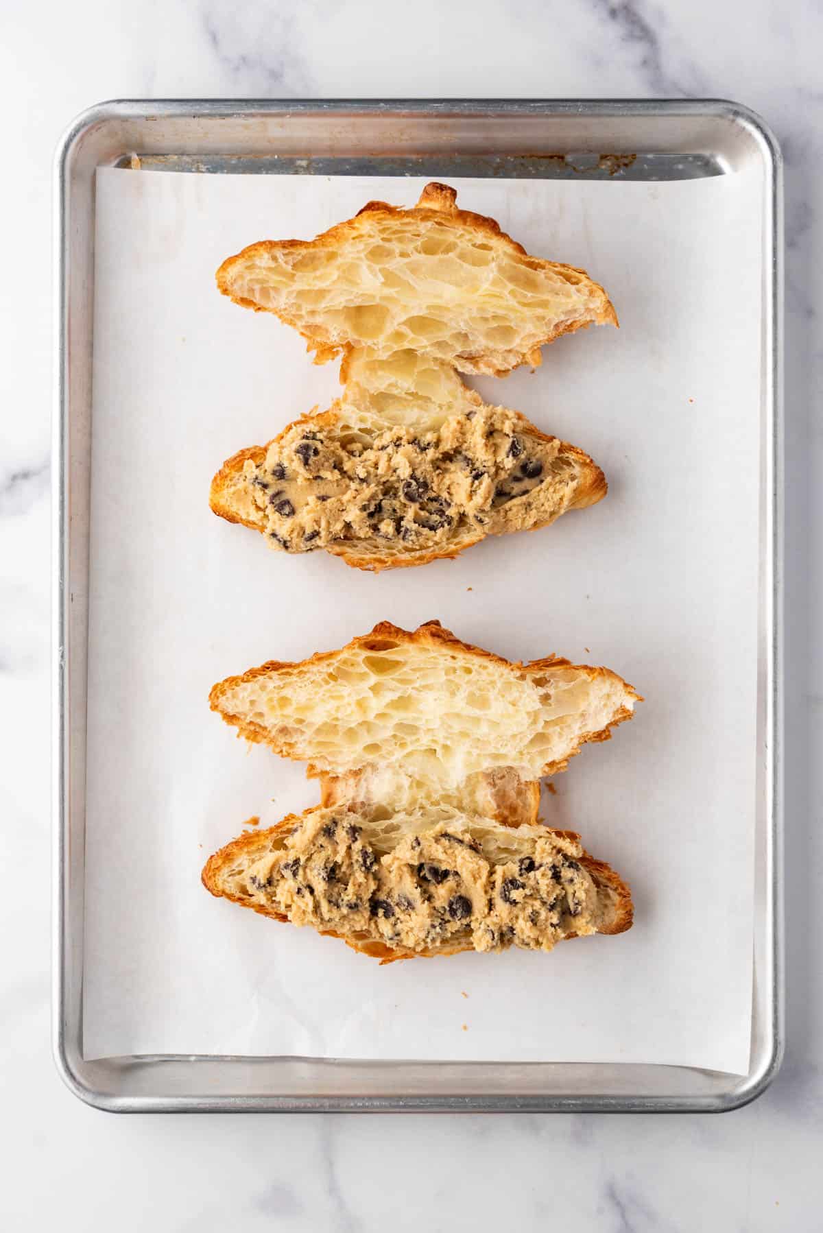 Croissants that have been sliced in half horizontally with chocolate chip cookie dough spread on the inside.