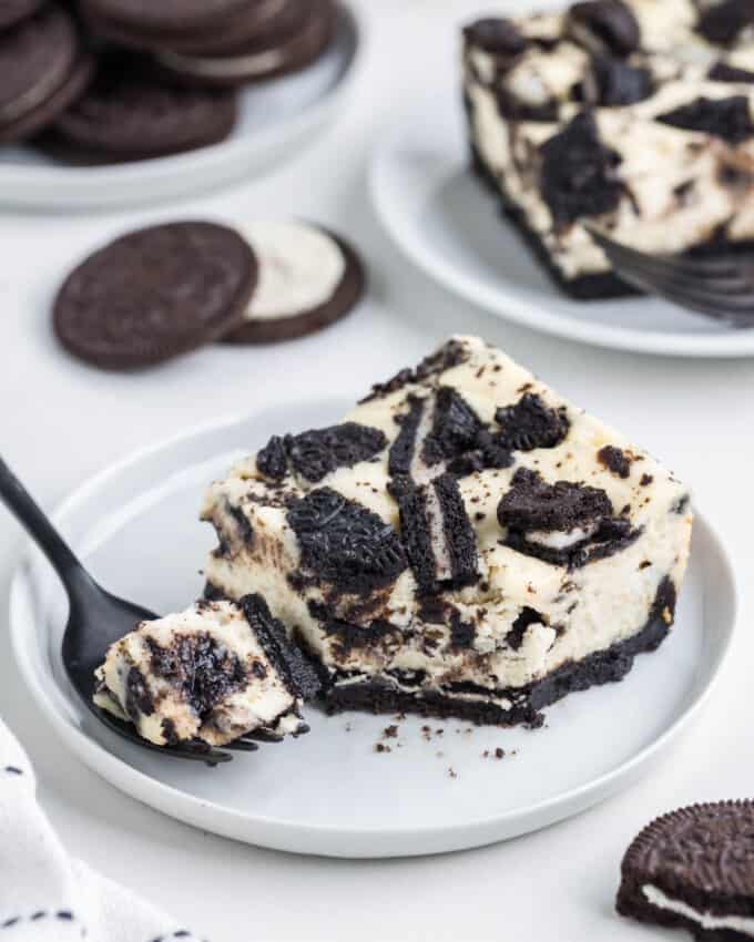 An Oreo cheesecake bar square on a white plate with a fork removing one bite from it with whole Oreos in the background.