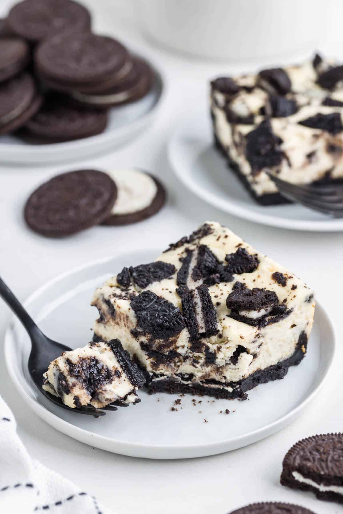 An Oreo cheesecake bar square on a white plate with a fork removing one bite from it with whole Oreos in the background.