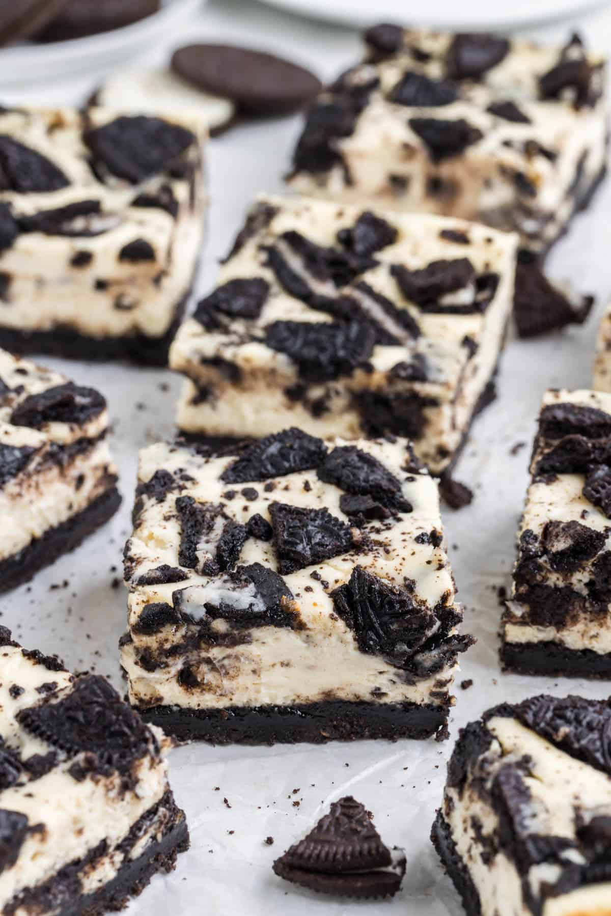 Oreo cheesecake squares scattered around a white parchment paper surface.