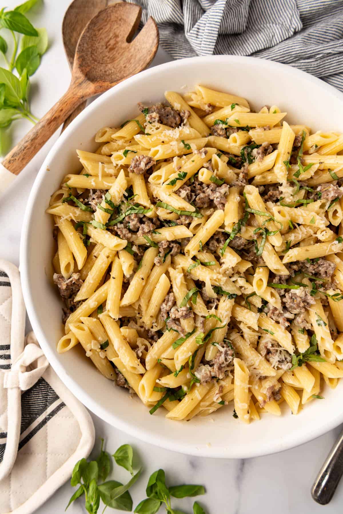 A serving bowl of penne pasta with Italian sausage next to serving utensils and fresh basil.