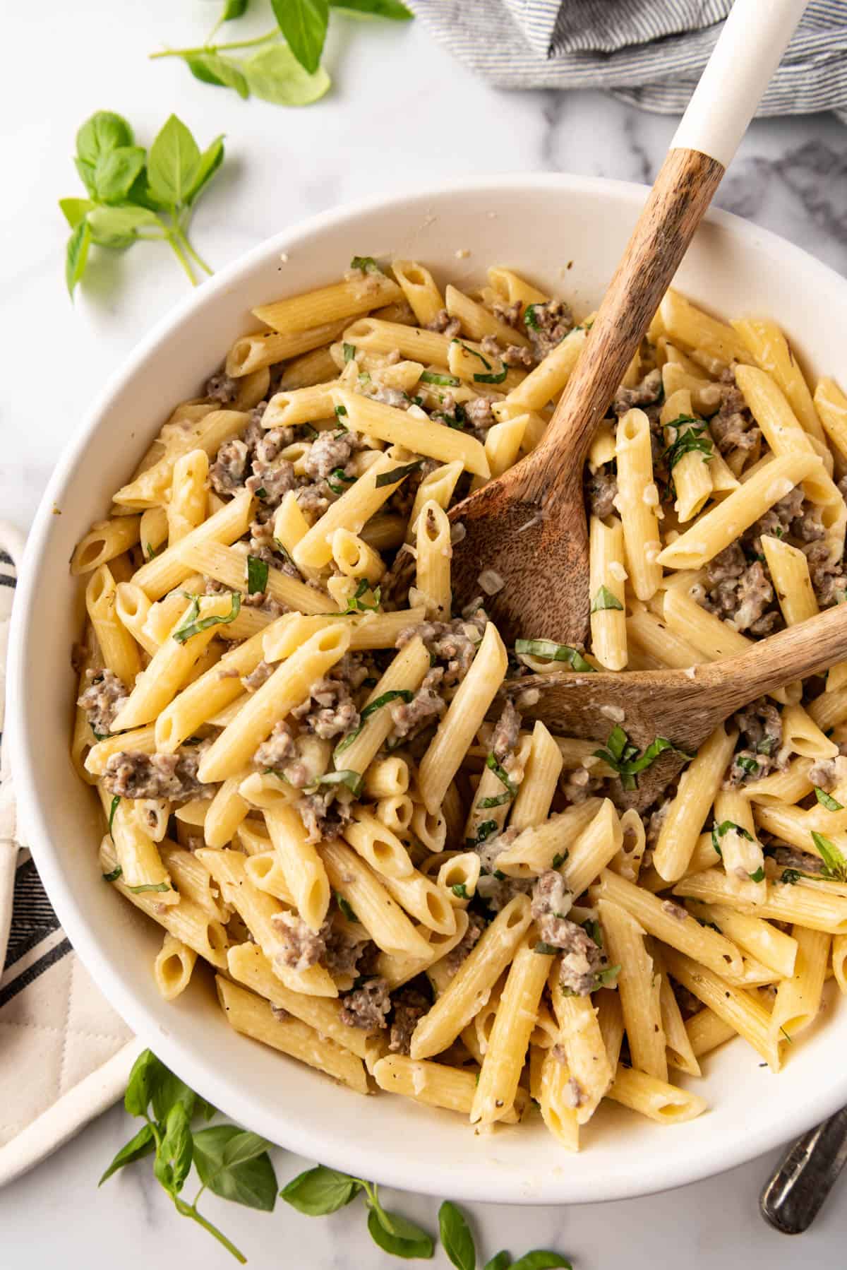 Serving spoons in a large white bowl of pasta with Italian sausage in a garlic cream sauce.