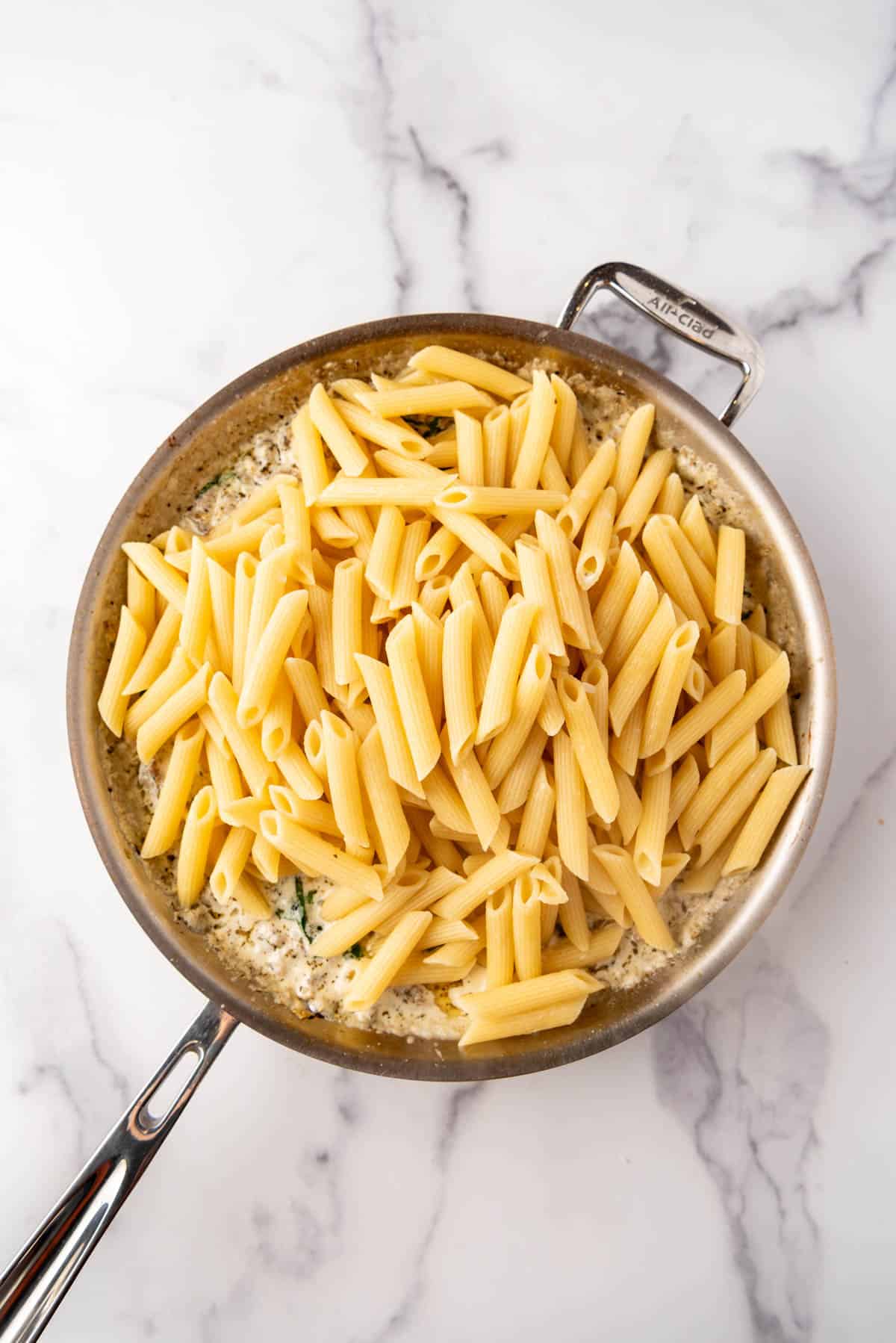 Adding cooked and drained penne pasta to a pan of cooked Italian sausage in creamy garlic sauce.