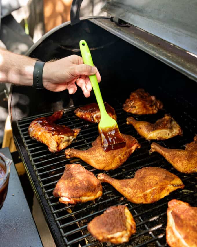 A hand holding a green silicon basting brush brushing barbecue sauce on smoked chicken on the Traeger.