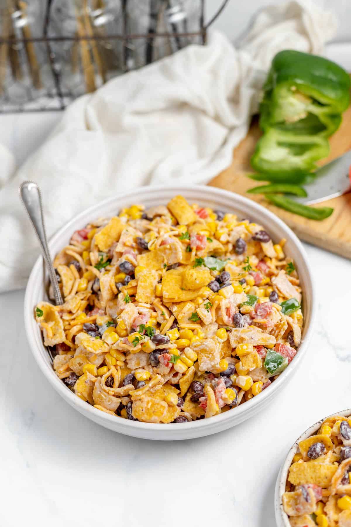 A large serving bowl of Frito corn salad in front of a cutting board with a green bell pepper on it.