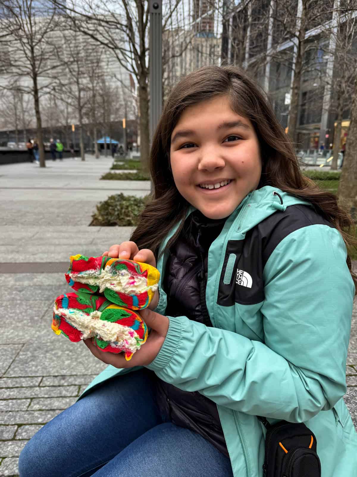 A girl holding a rainbow bagel with birthday cake cream cheese.