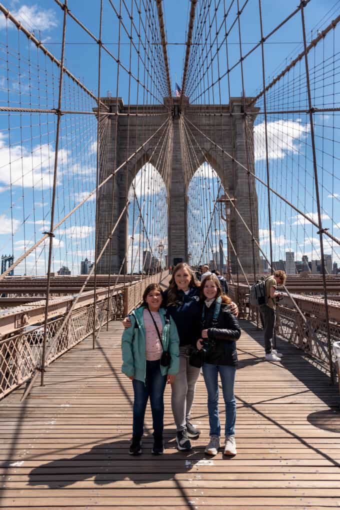 A mom and two daughters on the Brooklyn Bridge with the cables, tower, and blue sky behind them.