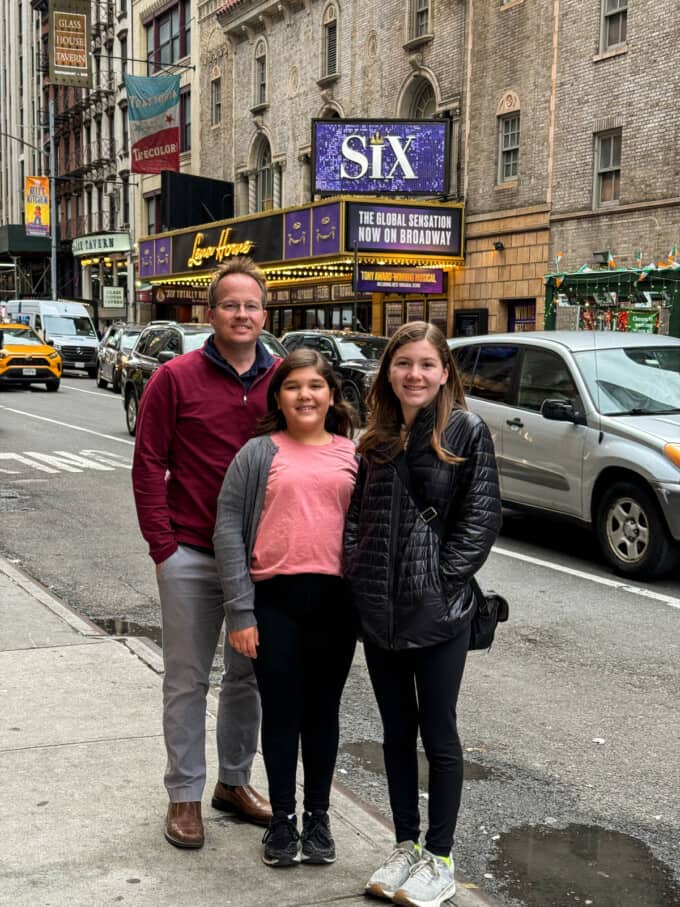 A dad and two daughters in front of the marquee for the musical Six on Broadway.