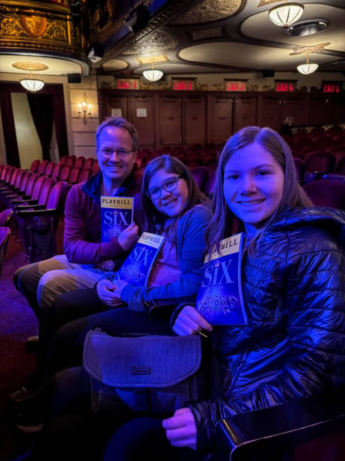 A dad and two daughters holding playbills for the musical Six in the seats in the theater.