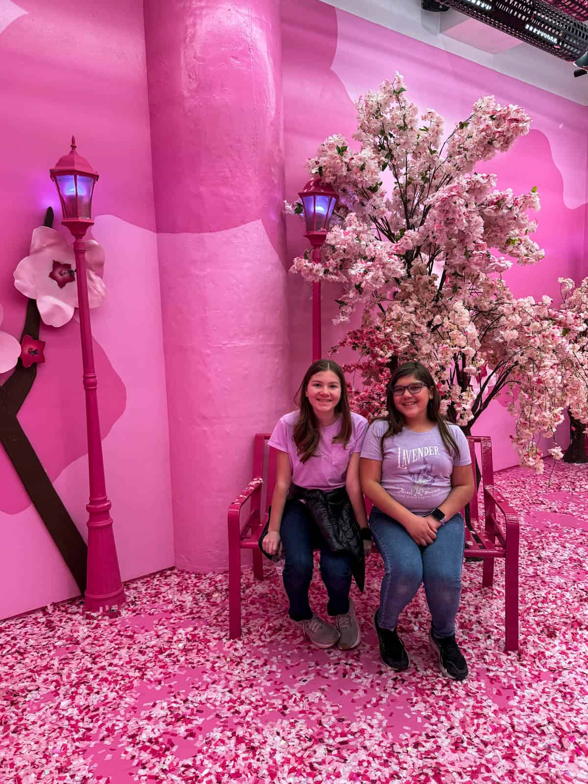 Two girls on a pink bench in a pink room by pink lamp posts in front of a pink tree.