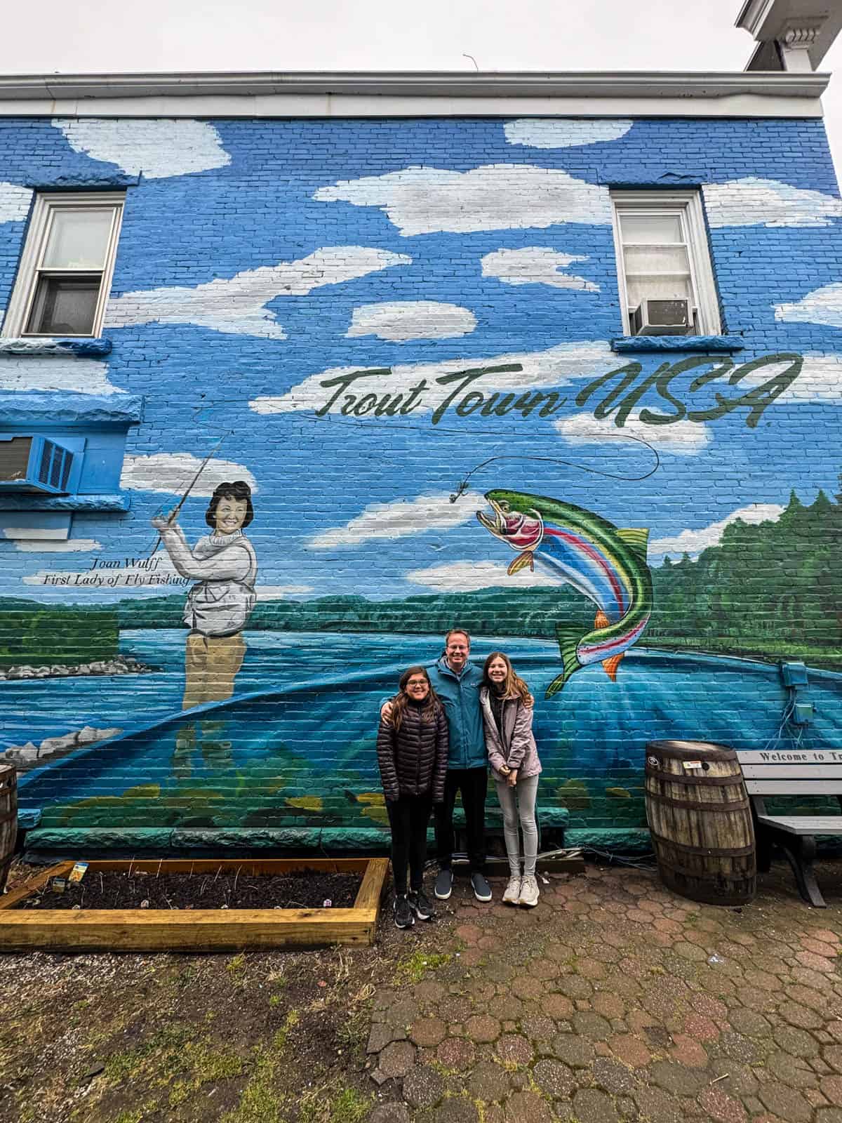 A dad and two daughters in front of a Trout Town, USA mural.