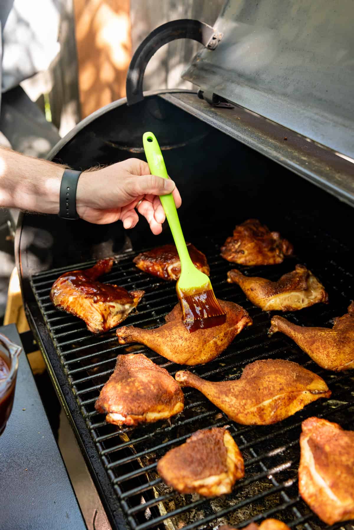 A hand holding a green silicon basting brush brushing barbecue sauce on smoked chicken on the Traeger.