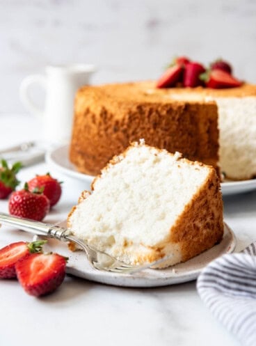 A slice of angel food cake on a white plate next to a striped cloth napkin and strawberries.