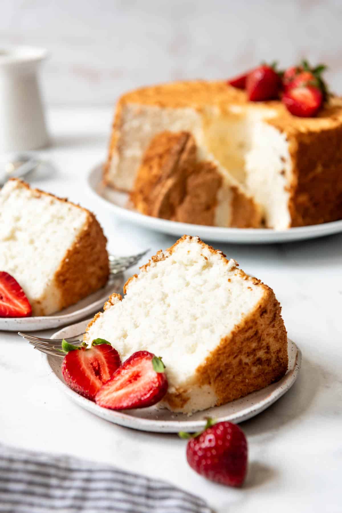 A slice of angel food cake on a plate in front of the rest of the cake.