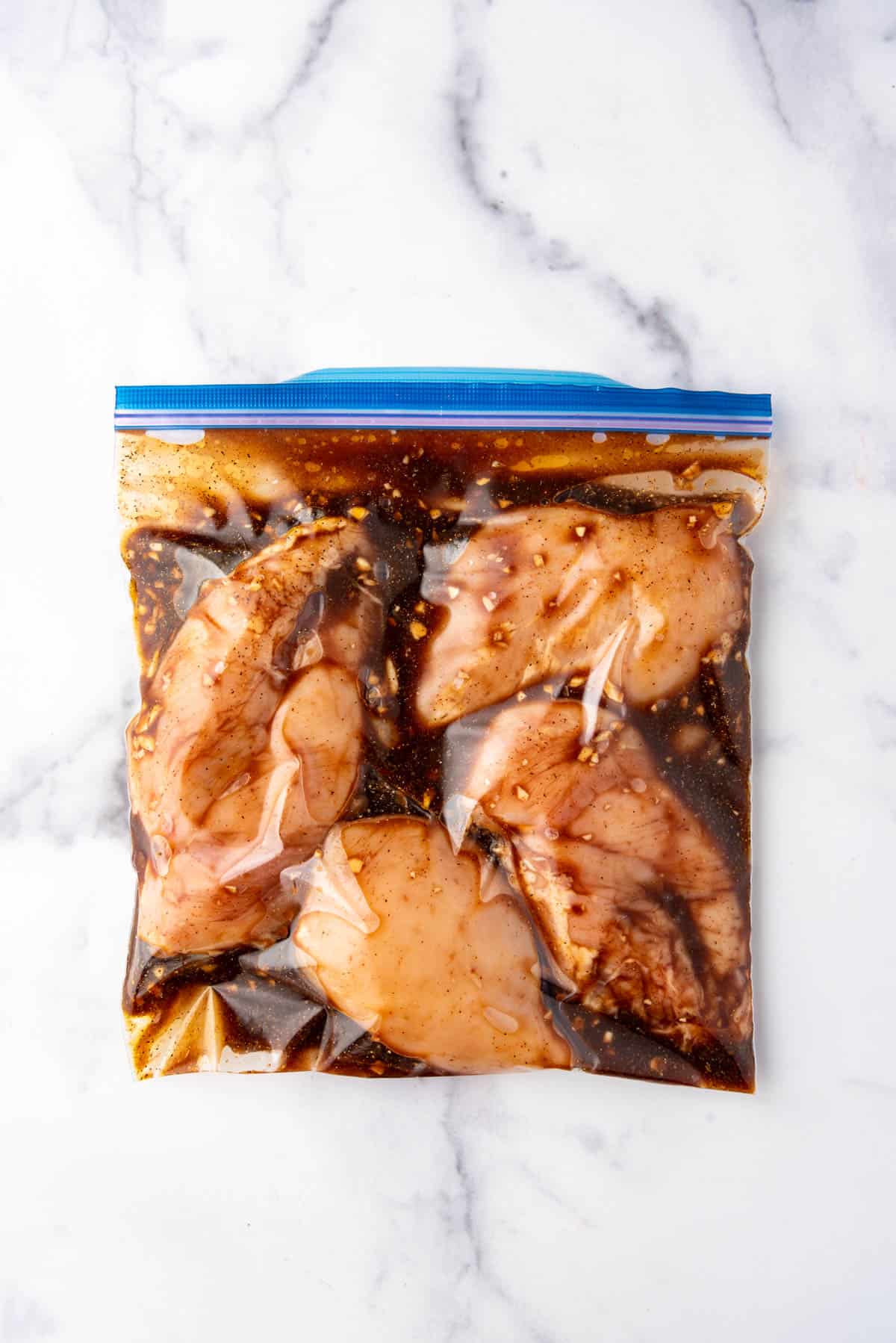 A ziploc bag full of chicken in a marinade on a white marble surface.