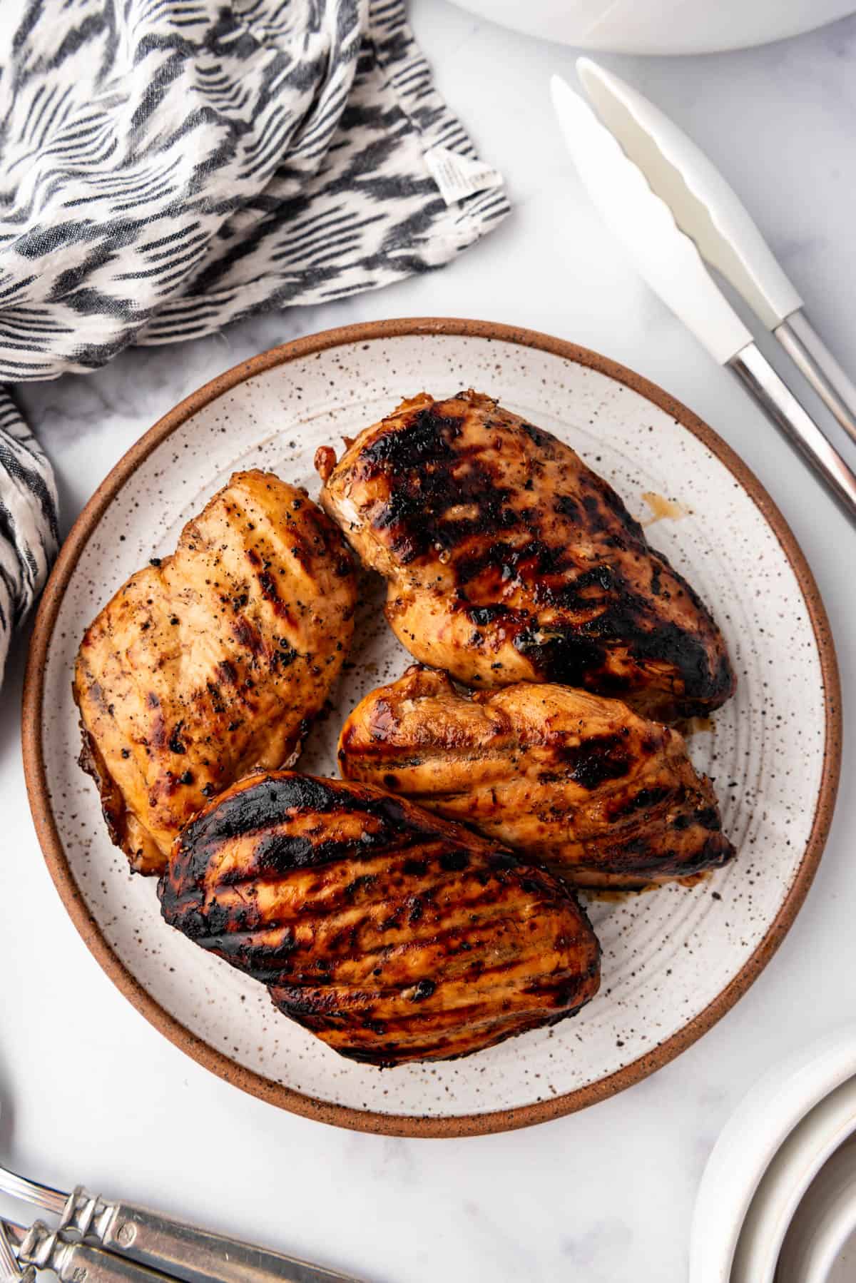 An overhead image of grilled marinated chicken breasts on a plate next to white tongs and a cloth napkin.