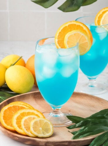 Two blue lagoon mocktails on a wooden plate with sliced citrus fruit nearby.