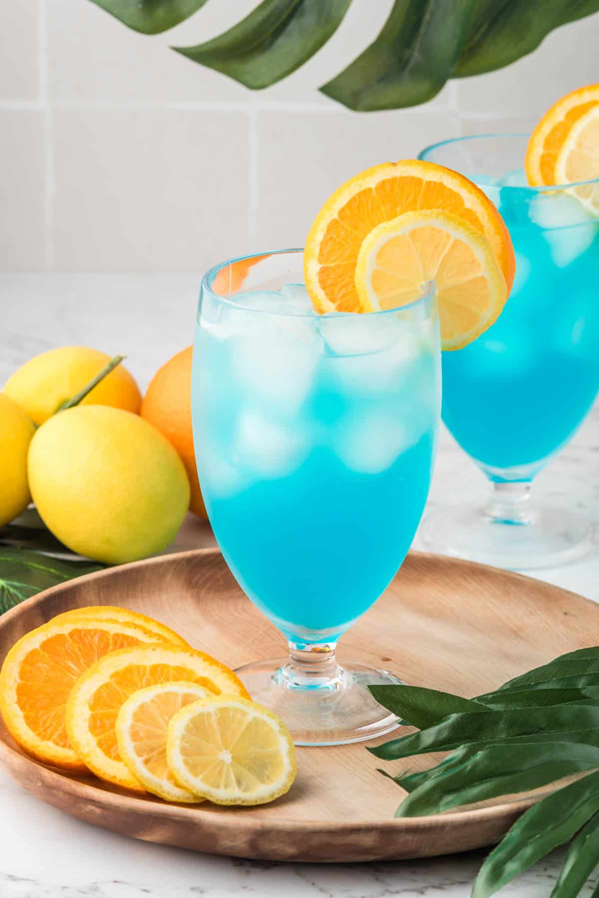 Two blue lagoon mocktails on a wooden plate with sliced citrus fruit nearby.