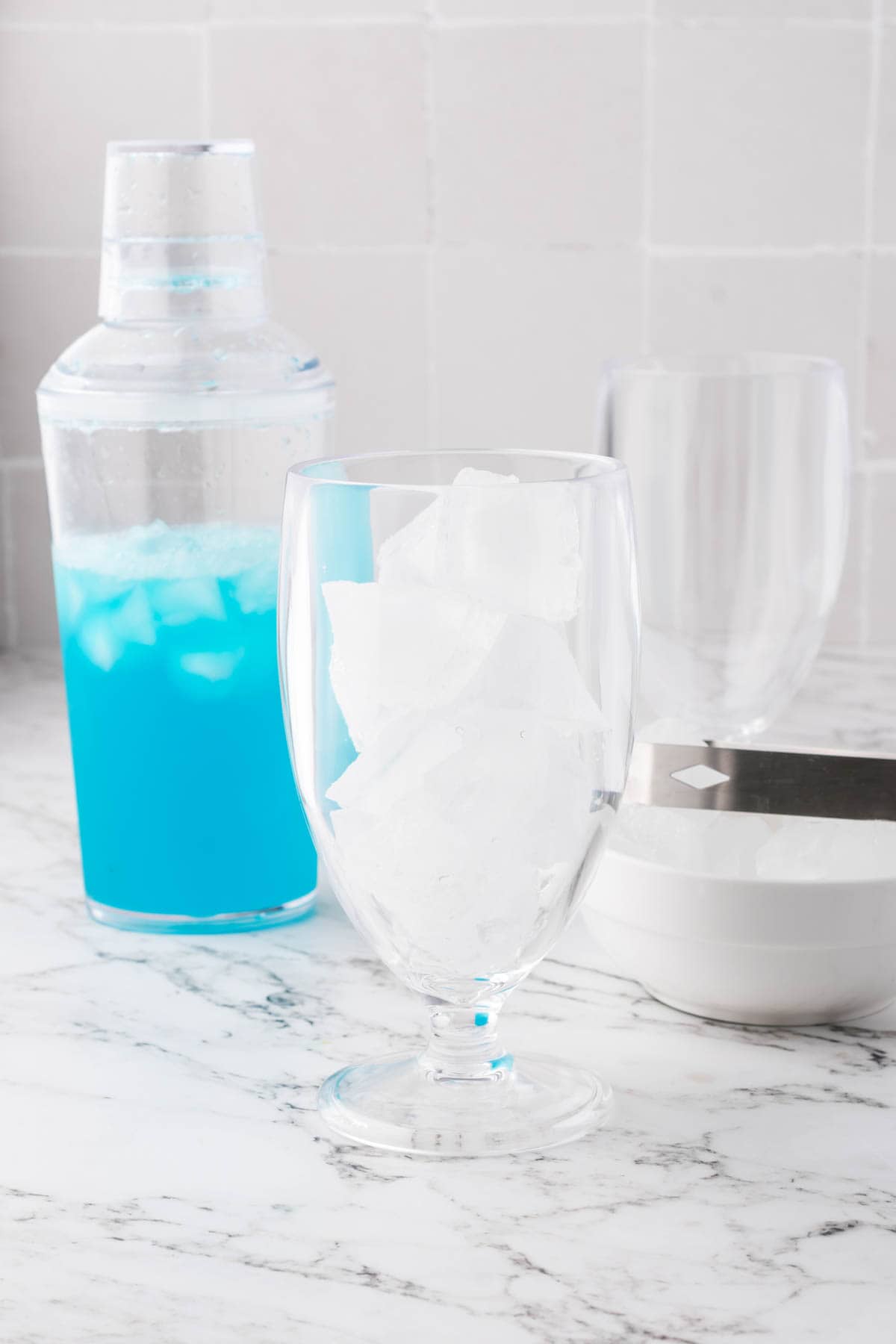 A glass full of ice cubes in front of a shaker of blue lagoon mocktail.