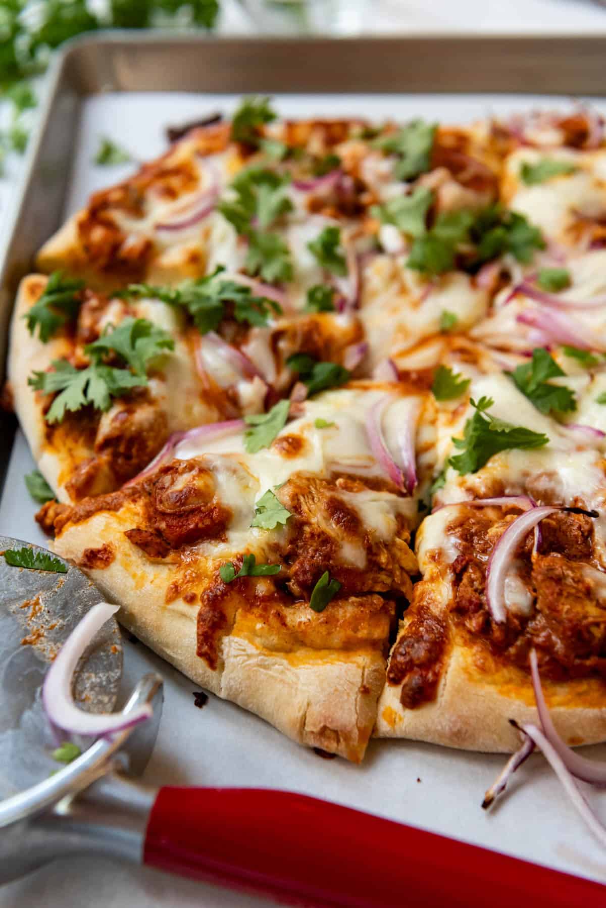 A side view of a chicken tikka masala pizza on a baking sheet lined with parchment paper next to a pizza cutter.