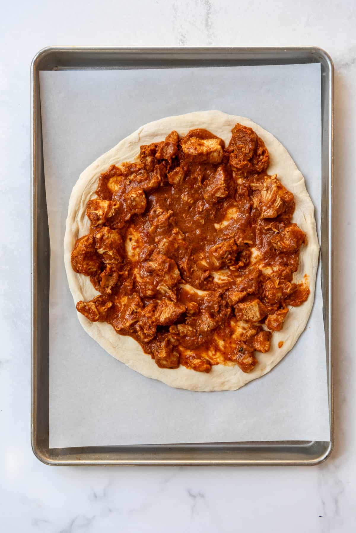 Spreading chicken tikka masala onto an unbaked pizza crust on a baking sheet.