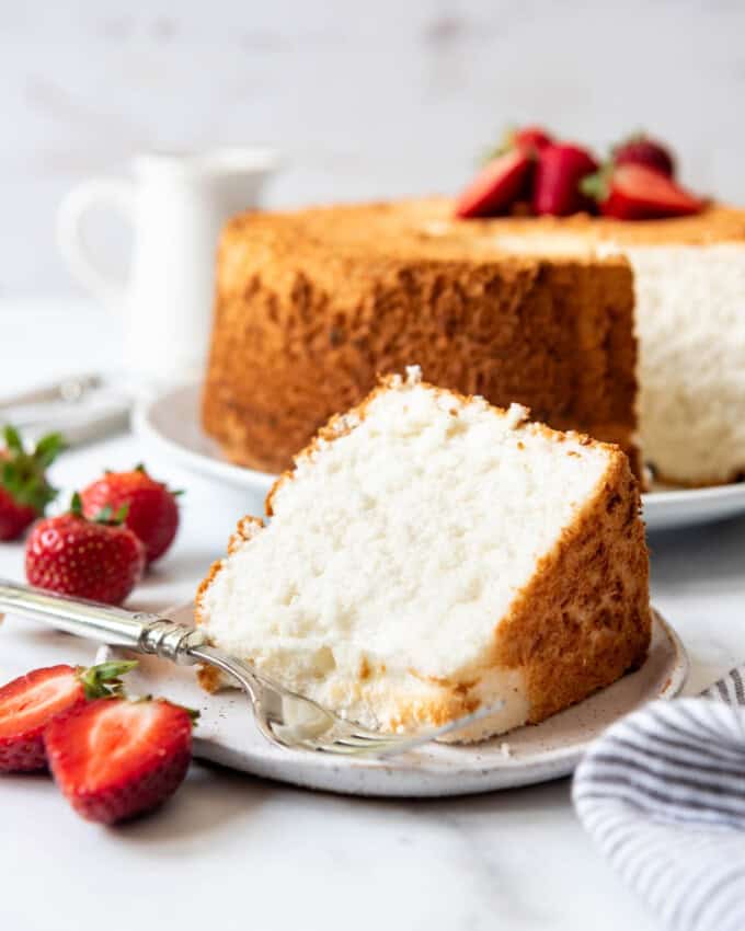 A slice of angel food cake on a white plate next to a striped cloth napkin and strawberries.