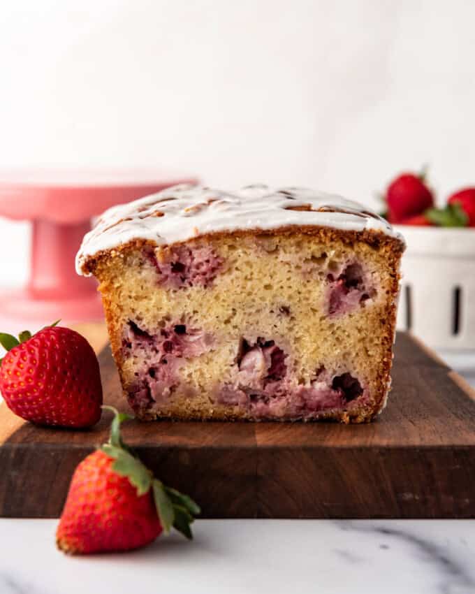 An image of a loaf of strawberry bread on a wooden cutting board with strawberries next to it.