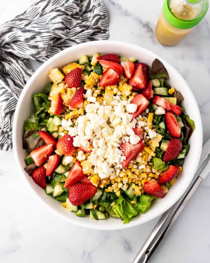 Adding sliced strawberries, grilled corn kernels, and feta cheese to a large salad bowl.