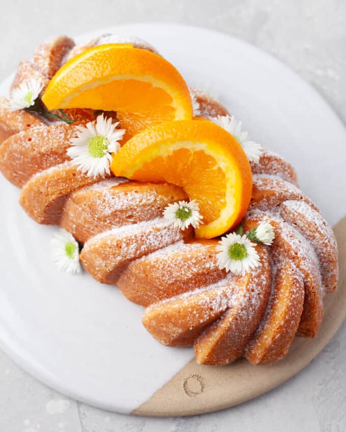 Overhead view of Orange Loaf Cake on a white serving plate with slices of orange and small white flowers garnishing it.