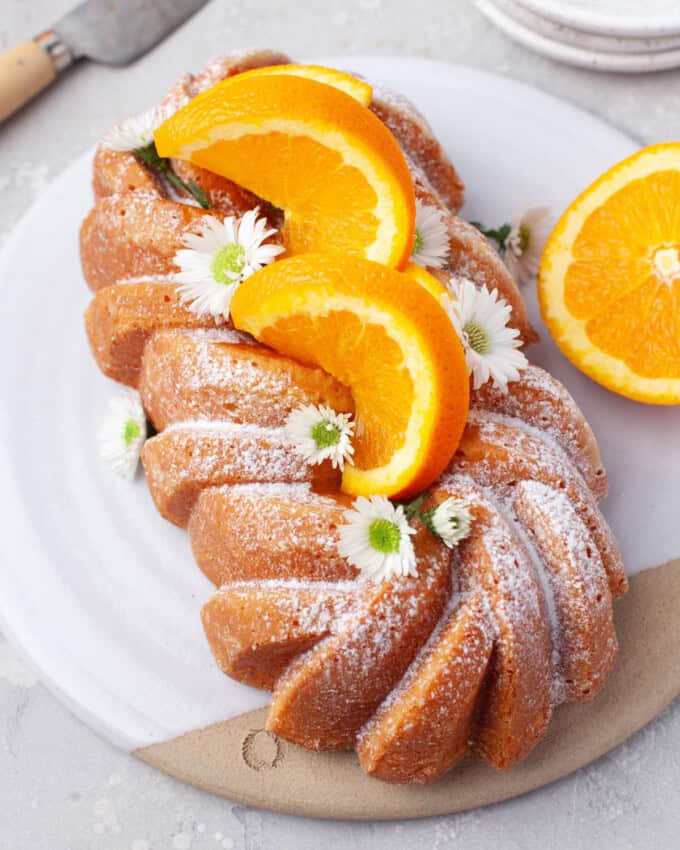 Overhead view of Orange Loaf Cake on a white serving plate with slices of orange and small white flowers garnishing it.