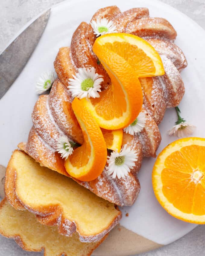 Overhead view of Orange Loaf Cake on a white serving plate with slices of orange and small white flowers garnishing it.