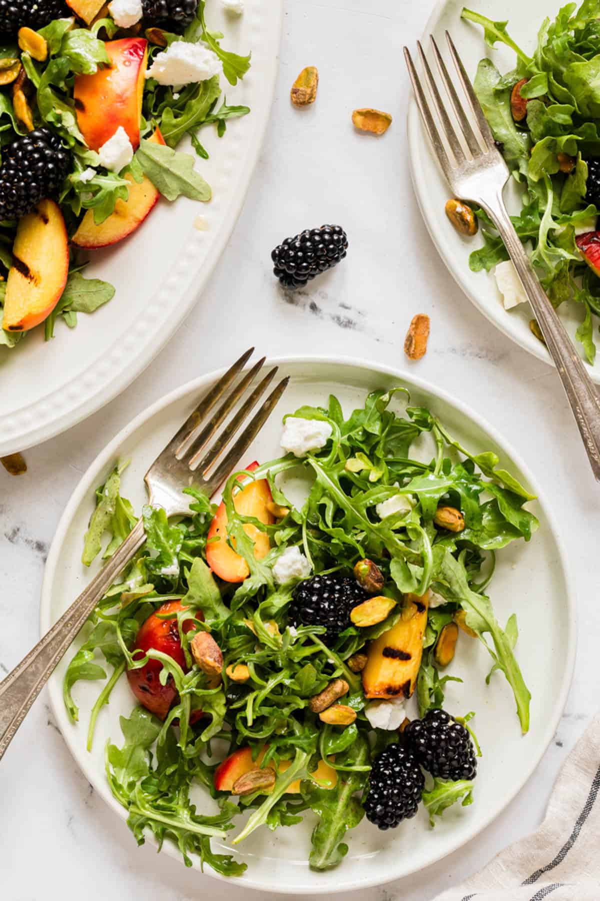 An overhead image of plates of arugula peach salad with scattered blackberries and pistachios around.