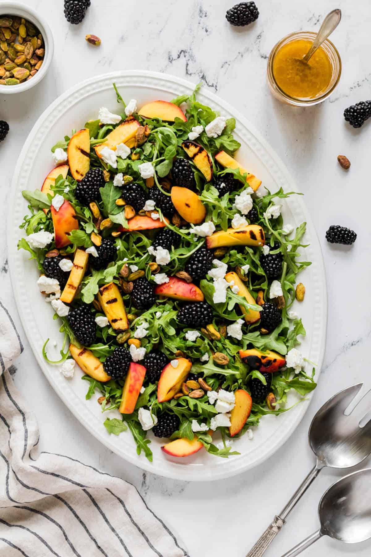 An overhead image of a grilled peach salad surrounded by napkins, dressing, blackberries, and serving spoons.