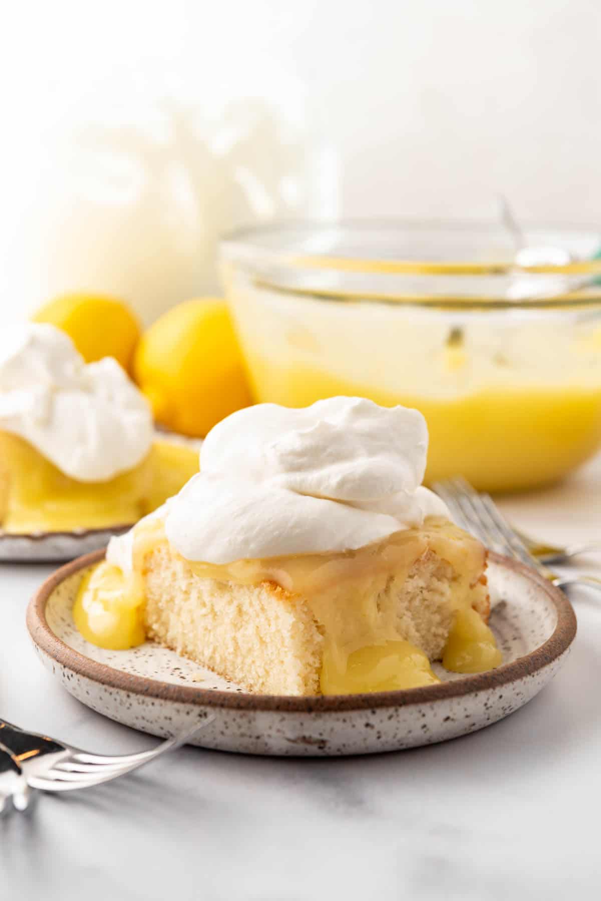 A close image of a piece of white cake with lemon topping and whipped cream in front of a large bowl of lemon pudding.