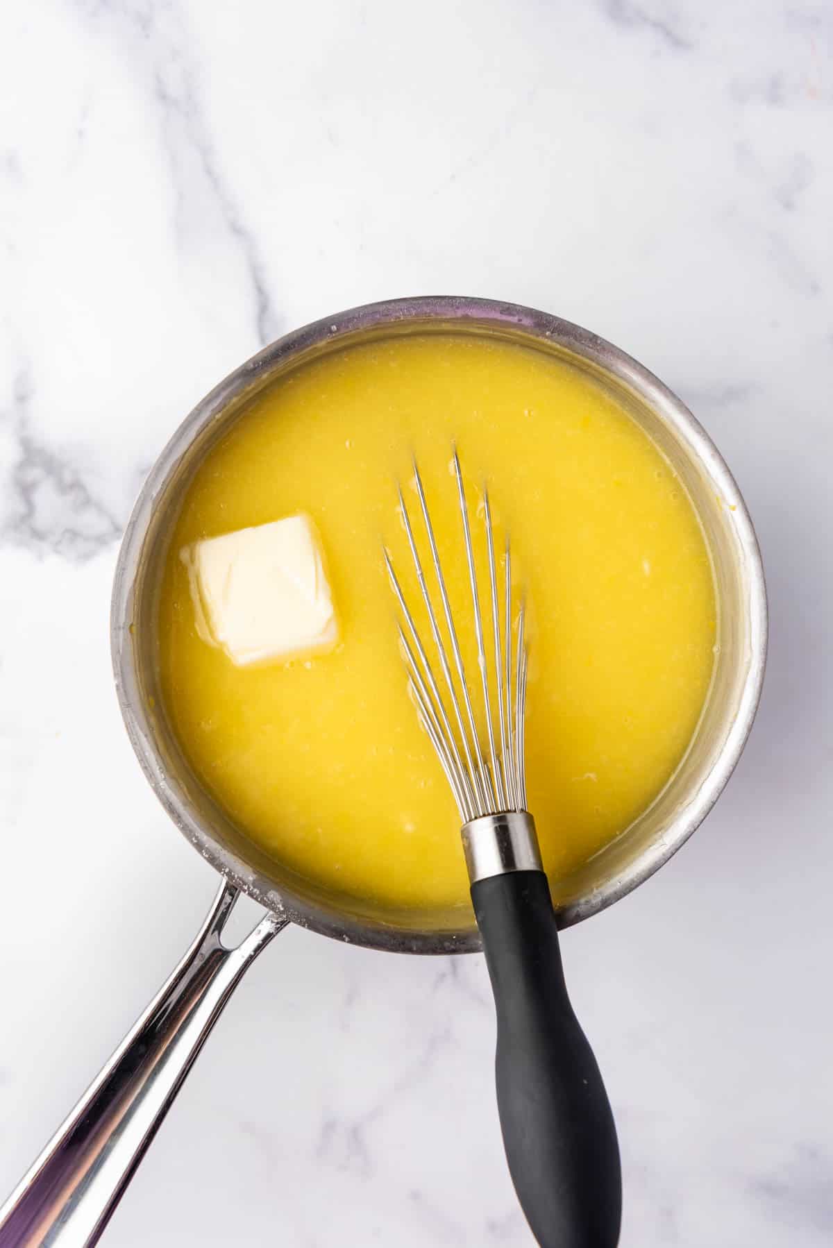 Adding butter to hot lemon pudding in a saucepan with a whisk.