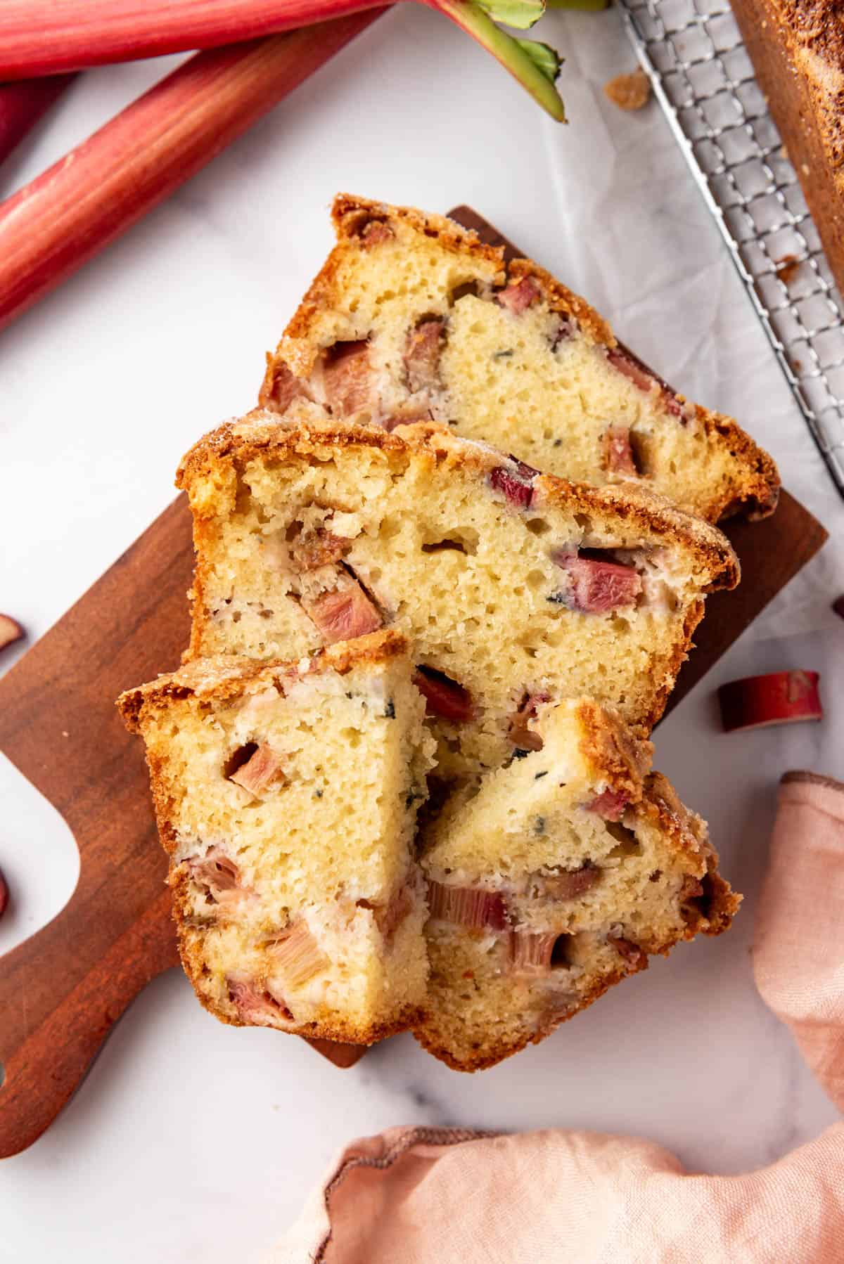 Slices of rhubarb quick bread on a wooden cutting board with stalks of rhubarb and a pink cloth napkin nearby.