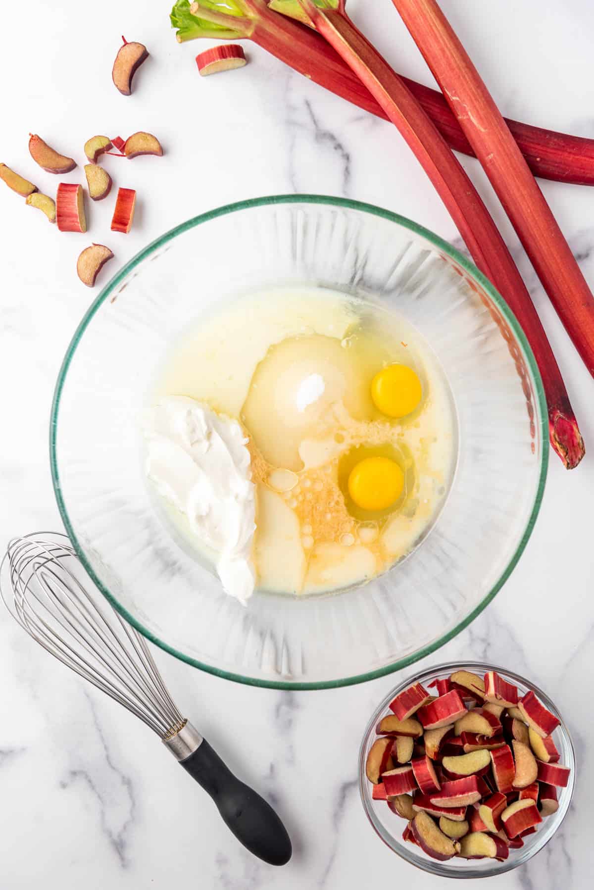 Combining wet ingredients in a large glass mixing bowl surrounded by rhubarb and a whisk.