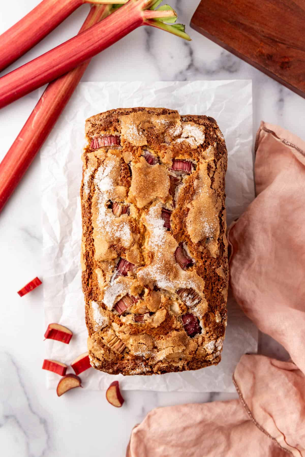 An overhead image of a loaf of rhubarb bread with a few stalks of pink rhubarb and a cloth napkin nearby.