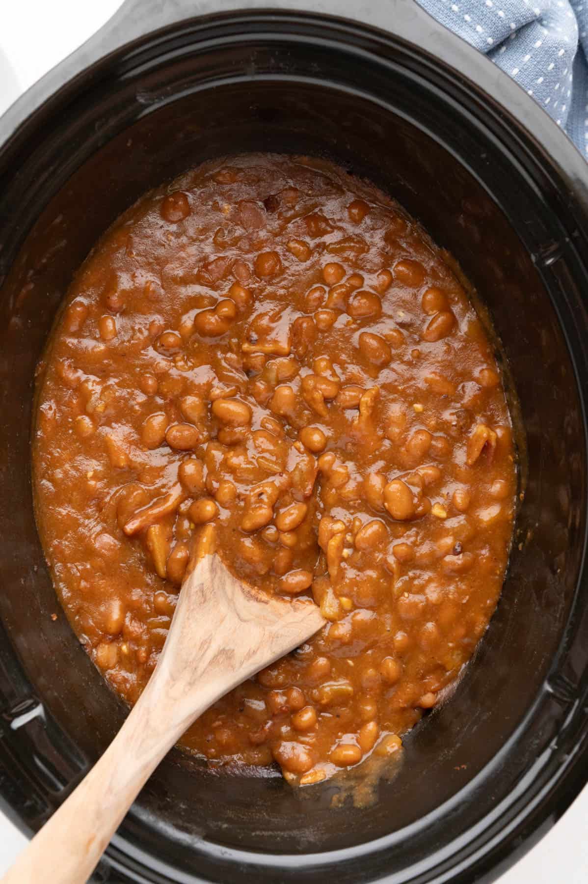 An overhead image of a pot of slow cooker baked beans with a wooden spoon in it.