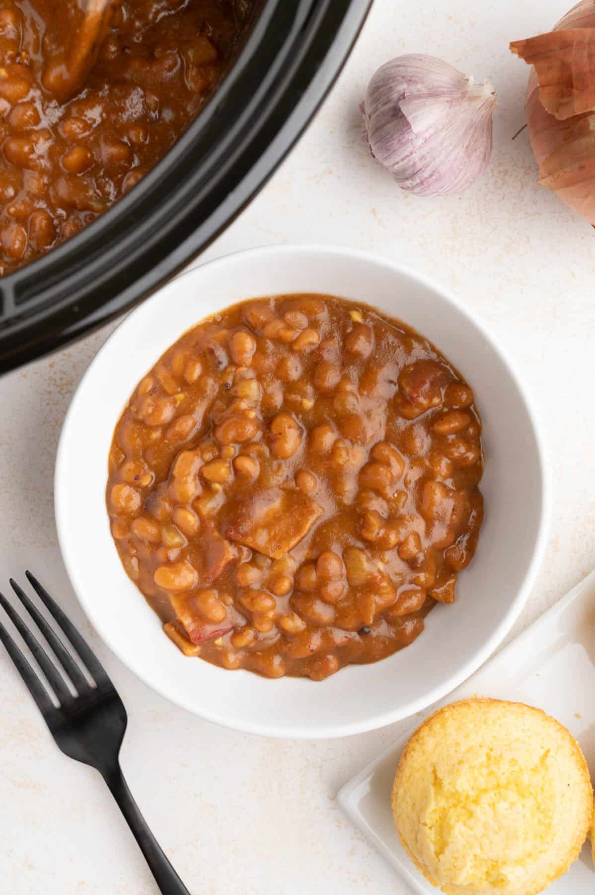 A bowl of baked beans next to a corn muffin and a slow cooker crock.