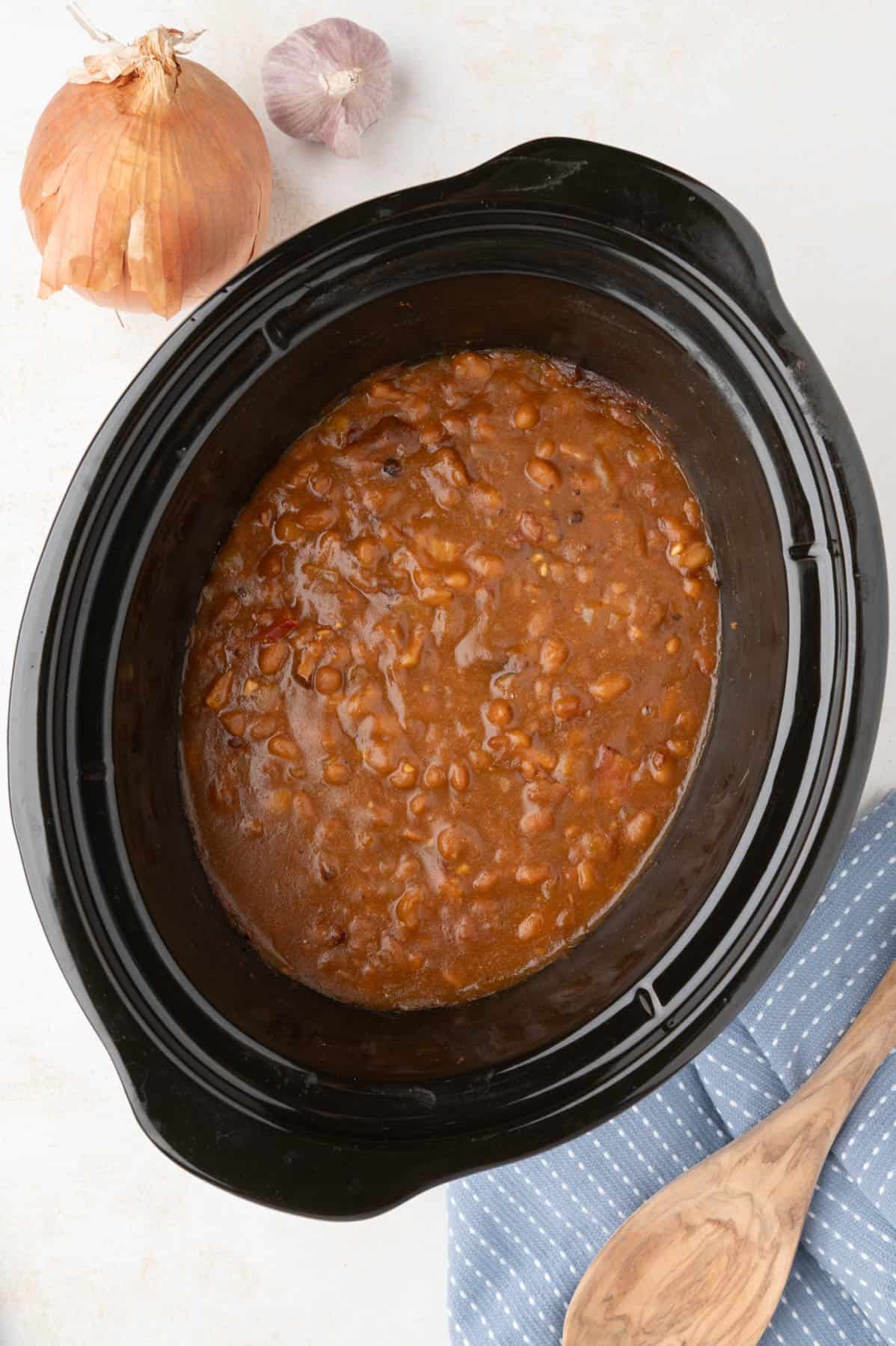 An overhead image of baked beans in a slow cooker next to a wooden spoon, onion, and garlic.