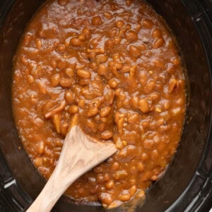 A close up image of slow cooker baked beans with a wooden spoon in them.