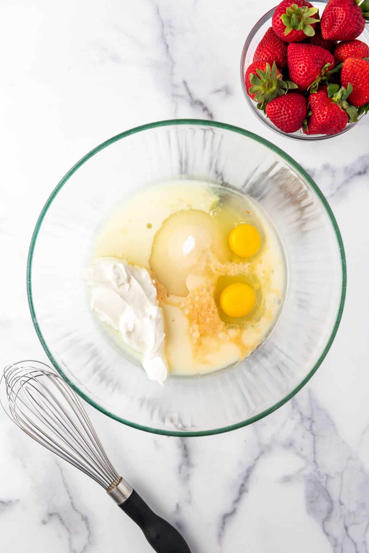 Adding wet ingredients to a bowl next to a whisk and a bowl of strawberries.