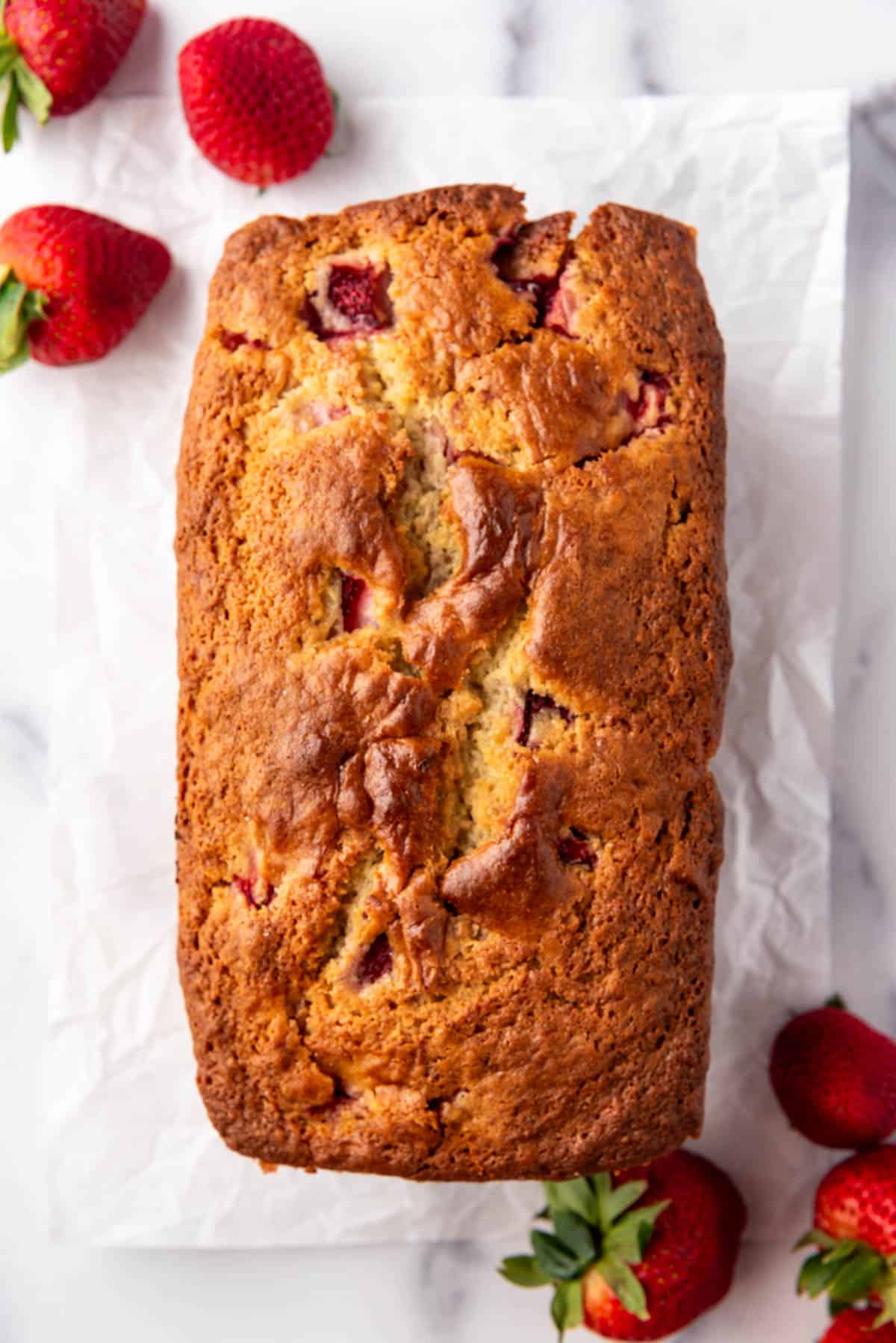 A baked loaf of strawberry bread on parchment paper with fresh strawberries scattered around.