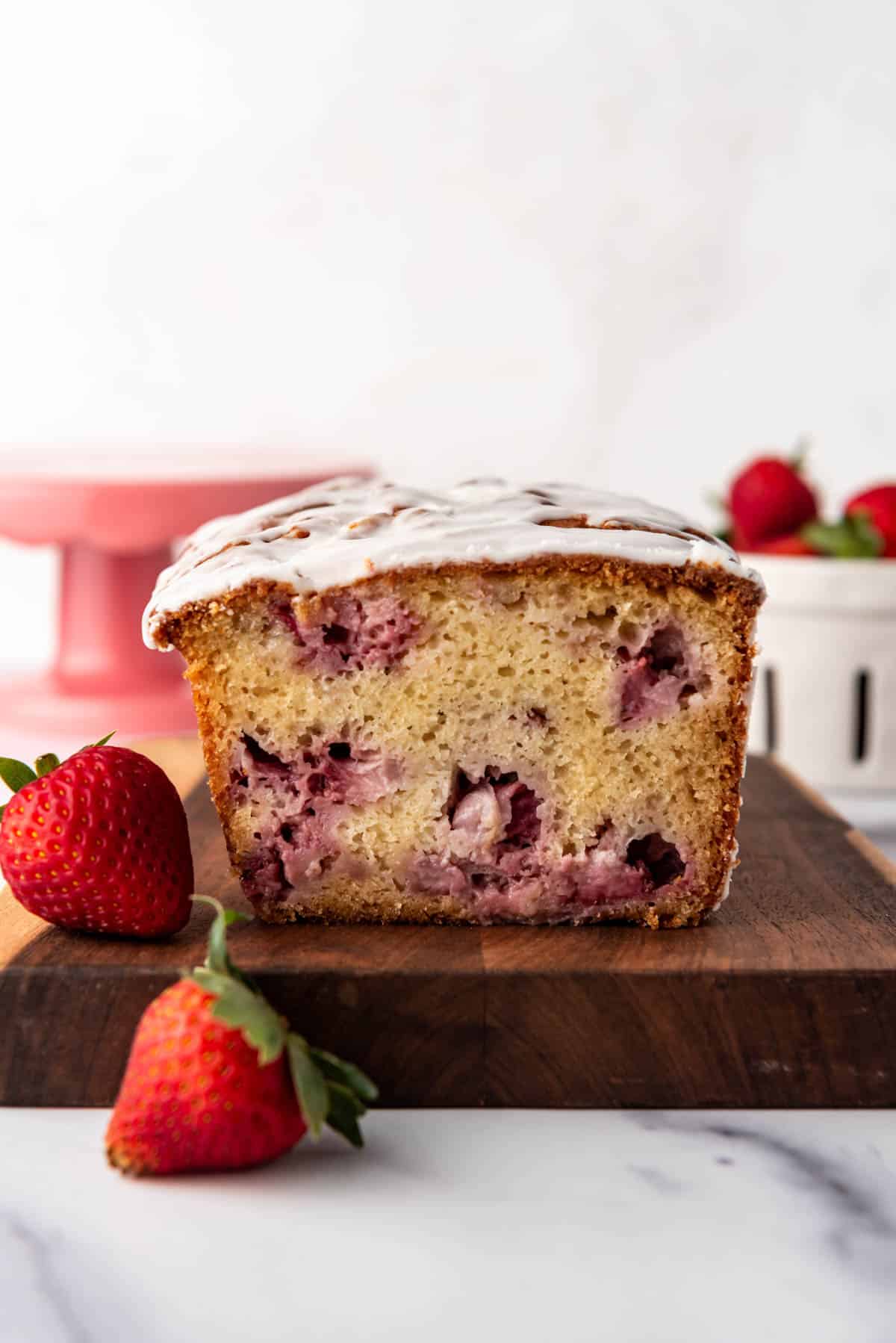 An image of a loaf of strawberry bread on a wooden cutting board with strawberries next to it.