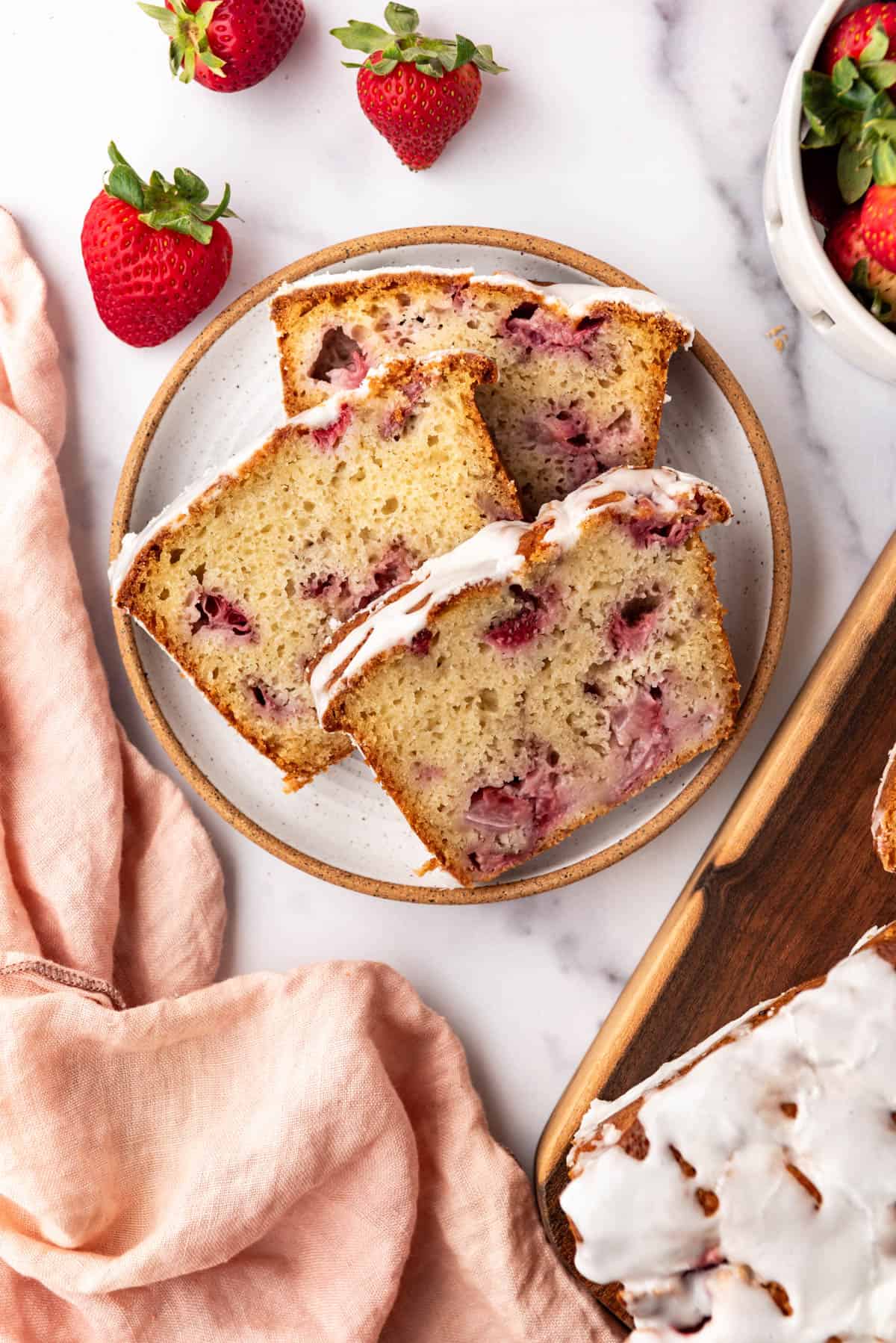 An overhead image of three slices of glazed strawberry bread on a plate with a pink cloth napkin and fresh strawberries beside it.