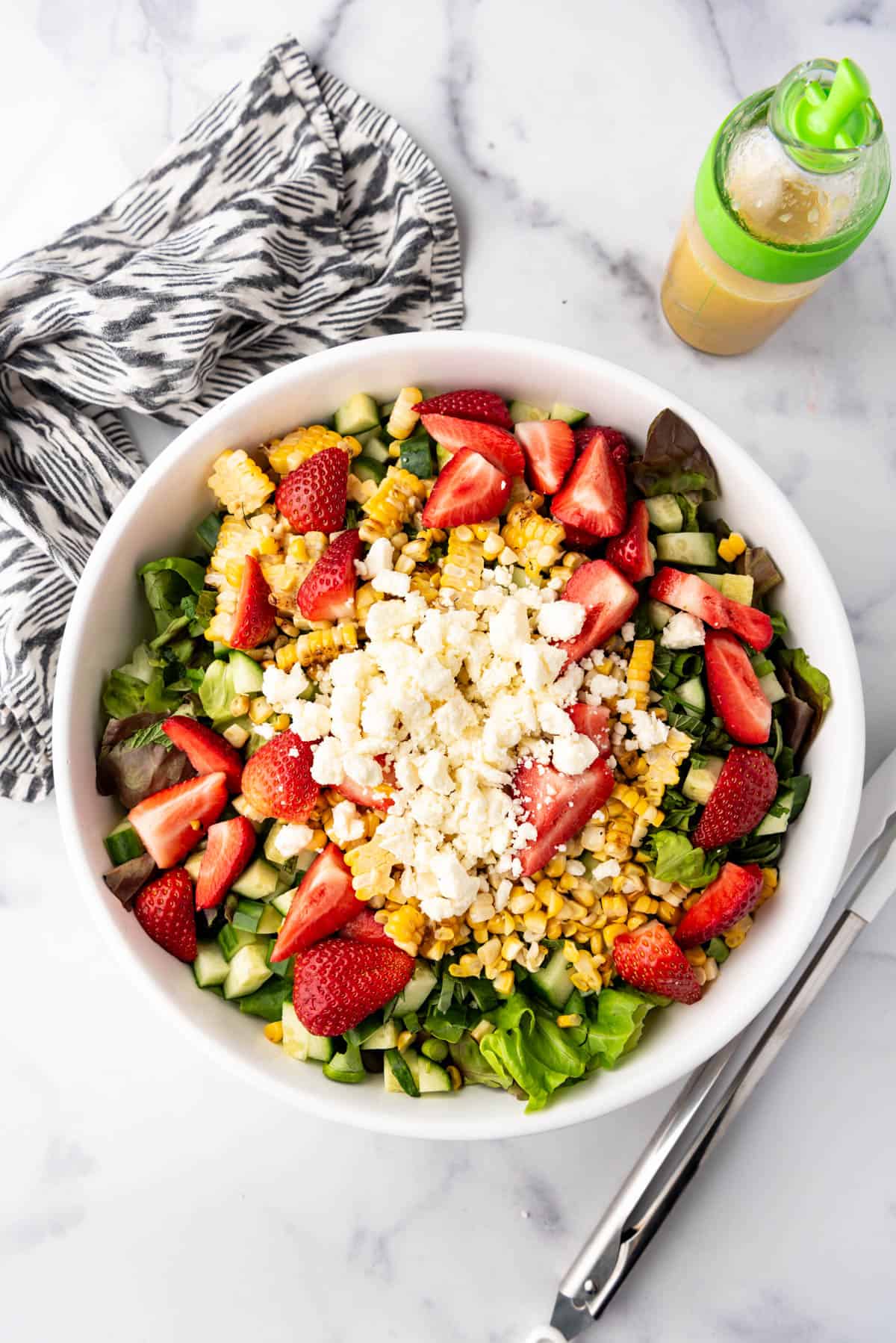 Adding sliced strawberries, grilled corn kernels, and feta cheese to a large salad bowl.