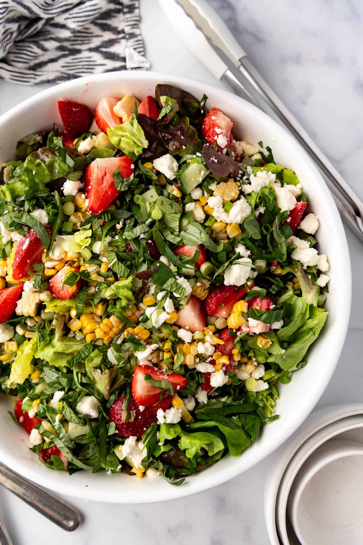 An overhead image of a strawberry corn summer salad in a large white serving bowl next to white tongs, bowls, and a linen napkin.